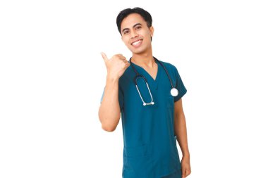 Mavi önlüklü, gülümseyen, kendine güvenen genç erkek hemşire, baş parmağıyla kenarı işaret ediyor, temiz beyaz bir arka planda izole edilmiş..