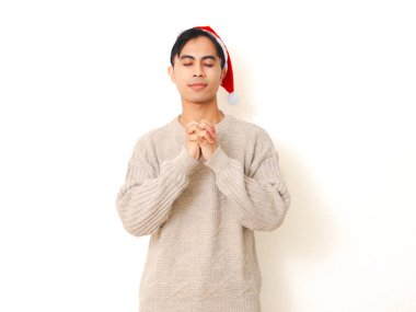 Gülümseyen Asyalı genç bir adam Noel Baba şapkası takıyor mutlu bir ifadeyle dua ediyor, beyaz arka planda izole edilmiş..