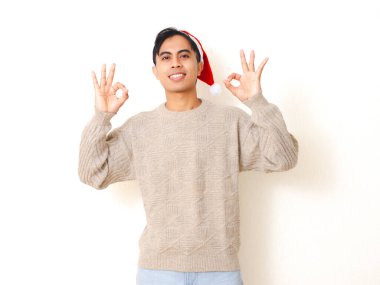 Noel Baba şapkası takan genç adam kendine güvenerek gülümsüyor ve temiz beyaz bir arkaplana karşı poz veren, bayram ve festival kavramları için mükemmel bir jest sergiliyor..