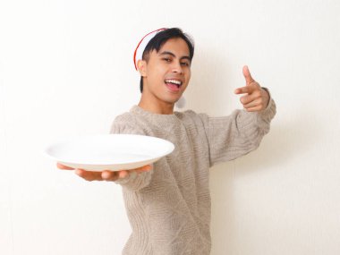 Heyecanlı genç Asyalı adam Noel Baba şapkası takıyor elinde fotokopi alanı olan boş bir tabak, Noel yemeği promosyonu ya da beyaz arka planda tatil reklamı için mükemmel..