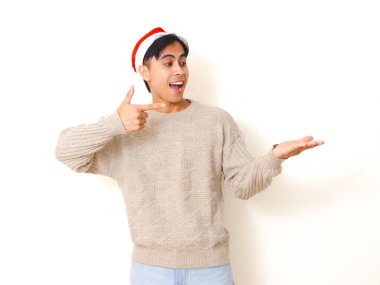 Noel Baba şapkası takan genç adam heyecanlı bir ifadeyle boş avucunu işaret ediyor, temiz beyaz bir arka planda izole edilmiş, tatil ve şenlik kavramları için mükemmel..