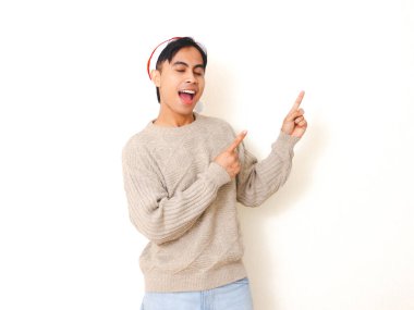 Noel şapkası takan genç adam şaşırtıcı bir ifadeyle kenarı işaret ediyor. Temiz beyaz bir arka plana karşı poz veriyor. Bayram ve tatil kavramları için mükemmel..