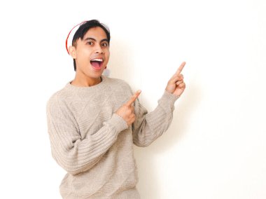 Noel şapkası takan genç adam şaşırtıcı bir ifadeyle kenarı işaret ediyor. Temiz beyaz bir arka plana karşı poz veriyor. Bayram ve tatil kavramları için mükemmel..