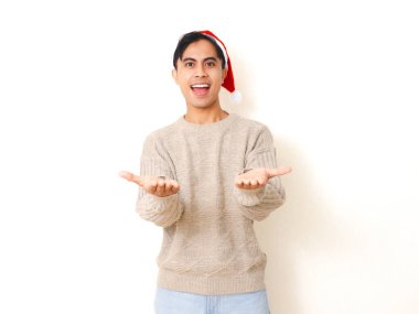 Noel Baba şapkası takan mutlu genç adam reklam konsepti için beyaz arka planda izole edilmiş ürünün tanıtımı için her iki avucunu da heyecanlı bir ifadeyle gösteriyor..