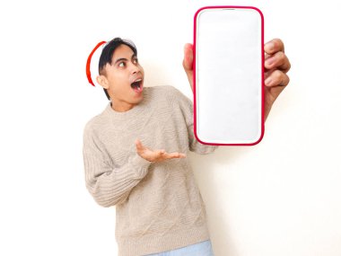 Genç Asyalı bir adam Noel şapkası takıyor. Beyaz izole edilmiş arka planda reklam ya da terfi için boş bir akıllı telefon model ekranı gösteriyor..