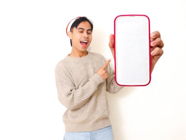 Noel şapkası takan genç Asyalı adam beyaz arka planda izole edilmiş, reklam ve tanıtım için boş akıllı telefon modelleme ekranını işaret ediyor..