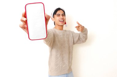 Noel şapkası takan genç Asyalı adam beyaz arka planda izole edilmiş, reklam ve tanıtım için boş akıllı telefon modelleme ekranını işaret ediyor..