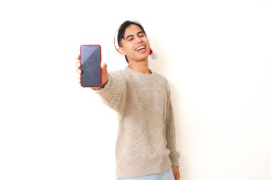Genç Asyalı bir adam Noel şapkası takıyor. Beyaz izole edilmiş arka planda reklam ya da terfi için boş bir akıllı telefon model ekranı gösteriyor..