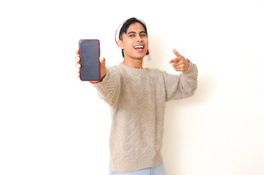 Noel şapkası takan genç Asyalı adam beyaz arka planda izole edilmiş, reklam ve tanıtım için boş akıllı telefon modelleme ekranını işaret ediyor..