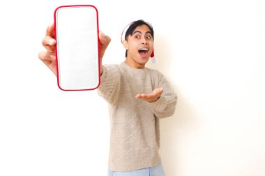 Genç Asyalı bir adam Noel şapkası takıyor. Beyaz izole edilmiş arka planda reklam ya da terfi için boş bir akıllı telefon model ekranı gösteriyor..