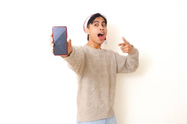 Noel şapkası takan genç Asyalı adam beyaz arka planda izole edilmiş, reklam ve tanıtım için boş akıllı telefon modelleme ekranını işaret ediyor..