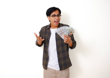 Şaşırmış, Asyalı genç bir adam elinde nakit para tutuyor, şok olmuş görünüyor, beyaz bir arka planda duruyor, beklenmedik bir para karşısında şaşkınlığını ve heyecanını dile getiriyor..