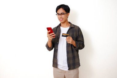 Gülümseyen Asyalı genç adam elinde bir ürün kartı tutarken beyaz arka planda telefonunu kullanıyor.