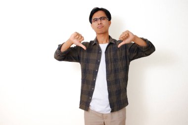 Asyalı ciddi bir adam, beyaz bir arka planda izole edilmiş kızgın bir ifadeyle baş parmağıyla bir jest yapıyor.