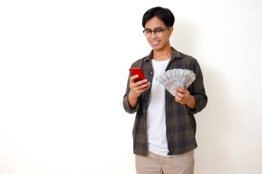 Mutlu Asyalı genç bir adamın portresi günlük kıyafetlerle cep telefonuna bakıyor, beyaz arka planda izole edilmiş para tutuyor.