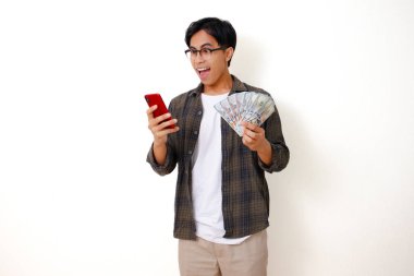 Mutlu Asyalı genç adam elinde para tutuyor ve telefonuna bakıyor. Heyecanlı ve şaşırmış bir ifadeyle, beyaz bir arka planda izole edilmiş.