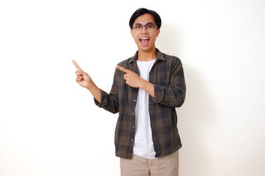 Heyecanlı genç Asyalı adam, izole edilmiş beyaz arka planda duruyor, gülümsüyor ve yanını işaret ediyor, neşeli bir ifade sergiliyor.