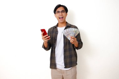 Mutlu Asyalı genç adam elinde para tutuyor ve telefonuna bakıyor. Heyecanlı ve şaşırmış bir ifadeyle, beyaz bir arka planda izole edilmiş.