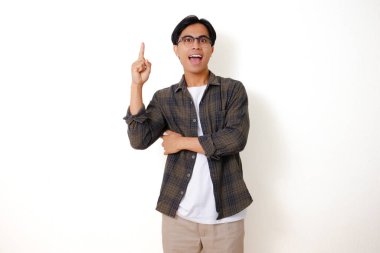 Heyecanlı genç Asyalı adam, beyaz bir arka planda izole edilmiş, parlak bir fikir ifadesi gösteren, düşünceli bir jestle yukarıya işaret ediyor.