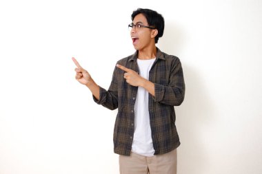 Heyecanlı genç Asyalı adam, izole edilmiş beyaz arka planda duruyor, gülümsüyor ve yanını işaret ediyor, neşeli bir ifade sergiliyor.