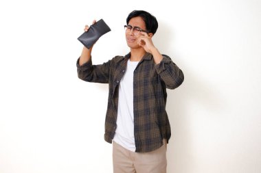 Asyalı üzgün genç bir adam, beyaz bir arka planda izole edilmiş, stresli bir ifadeyle boş bir cüzdanı tutuyor ve gösteriyor.