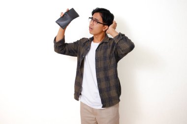 Asyalı üzgün genç bir adam, beyaz bir arka planda izole edilmiş, stresli bir ifadeyle boş bir cüzdanı tutuyor ve gösteriyor.