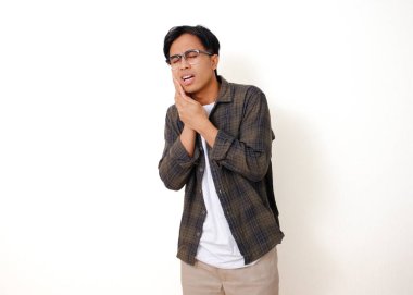 Sağlıksız genç Asyalı adam, beyaz arka planda izole edilmiş acı dolu bir ifadeyle yanağına dokunuyor.