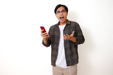 Mutlu, Asyalı genç bir adam, beyaz arka planda izole edilmiş, etkileyici bir yüz ifadesiyle cep telefonuna bakıyor.