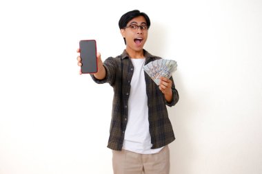 Heyecanlı genç Asyalı adam elinde para tutuyor ve boş bir cep telefonu ekranı gösteriyor, gülümsüyor, beyaz bir arka planda izole edilmiş.