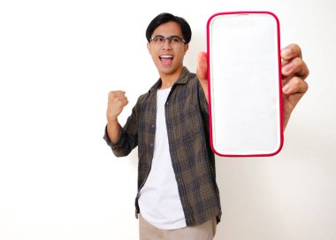 Heyecanlı genç Asyalı adam elinde boş bir cep telefonu ekranı ile başarıyı beyaz bir arka planda izole edilmiş neşeli bir hareketle kutluyor.