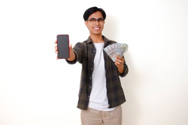 Heyecanlı genç Asyalı adam elinde para tutuyor ve boş bir cep telefonu ekranı gösteriyor, gülümsüyor, beyaz bir arka planda izole edilmiş.