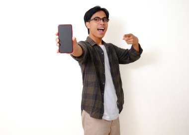 Heyecanlı genç Asyalı adam beyaz arka planda izole edilmiş boş bir cep telefonu ekranını işaret ediyor.