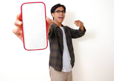 Heyecanlı genç Asyalı adam beyaz arka planda izole edilmiş boş bir cep telefonu ekranını işaret ediyor.