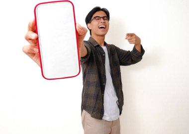 Heyecanlı genç Asyalı adam beyaz arka planda izole edilmiş boş bir cep telefonu ekranını işaret ediyor.