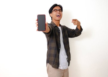 Heyecanlı genç Asyalı adam beyaz arka planda izole edilmiş boş bir cep telefonu ekranını işaret ediyor.