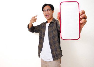 Heyecanlı genç Asyalı adam beyaz arka planda izole edilmiş boş bir cep telefonu ekranını işaret ediyor.