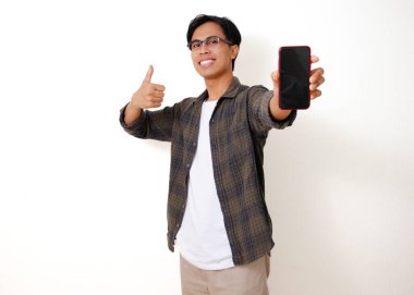 Heyecanlı bir Asyalı genç adam beyaz bir arka planda izole bir şekilde başparmak hareketi yaparken boş bir cep telefonu ekranı gösteriyor.