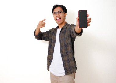Heyecanlı genç Asyalı adam beyaz arka planda izole edilmiş boş bir cep telefonu ekranını işaret ediyor.