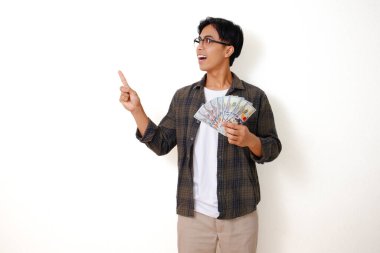 Beyaz arka planda izole edilmiş, işaret ederken elinde nakit para tutan mutlu Asyalı genç adam.