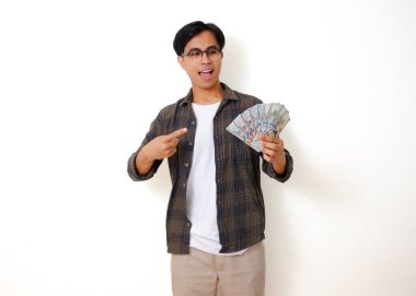 Beyaz arka planda izole edilmiş, işaret ederken elinde nakit para tutan mutlu Asyalı genç adam.