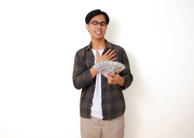 Gülümseyen Asyalı genç adam para tutuyor ve göğsüne rahatlamış bir ifadeyle dokunuyor, beyaz arka planda izole edilmiş.