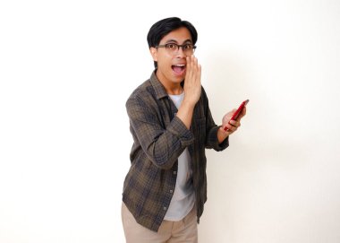 Beyaz arka planda izole edilmiş, fısıldayan bir jest yaparken akıllı telefon tutan mutlu Asyalı genç adam.