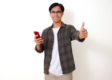 Akıllı telefon tutan gülümseyen Asyalı genç adam, temiz beyaz bir arka planda izole edilmiş, baş parmak işareti yapıyor.
