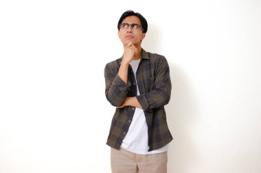 Ciddi bir Asyalı genç adam ayakta duruyor ve çenesini düşünceli bir ifadeyle tutuyor, izole edilmiş beyaz arka planda bir düşünce hareketi gösteriyor.