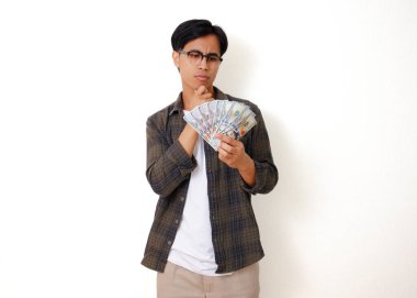 Stresli genç Asyalı adam para tutuyor ve kafası karışmış bir ifadeyle kafasını kaşıyor, beyaz arka planda soyutlanmış.