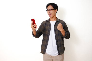 Heyecanlı Asyalı genç adam, cep telefonuna bakıp yumruğunu sıkarken, beyaz arka planda izole bir şekilde başarıyı kutluyor.