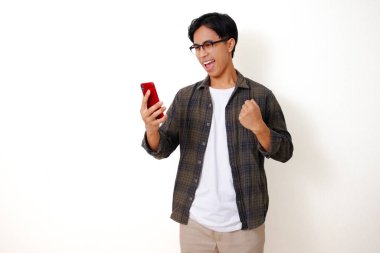 Heyecanlı Asyalı genç adam, cep telefonuna bakıp yumruğunu sıkarken, beyaz arka planda izole bir şekilde başarıyı kutluyor.