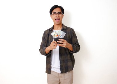 Gülümseyen Asyalı genç bir adam para dolu bir cüzdan taşıyor, mutluluk ve finansal başarı yayıyor. Temiz beyaz bir arkaplanda izole edilmiş.