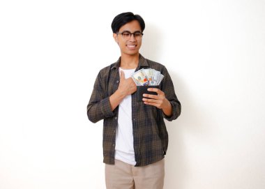 Gülümseyen Asyalı genç bir adam para dolu bir cüzdan taşıyor, mutluluk ve finansal başarı yayıyor. Temiz beyaz bir arkaplanda izole edilmiş.