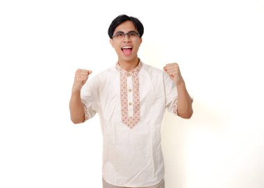 Heyecanlı Endonezyalı Müslüman adam sevinçli bir kutlama jesti ile iki elini kaldırıyor, beyaz arka planda izole edilmiş, başarıyı, mutluluğu ve pozitif enerjiyi simgeliyor..